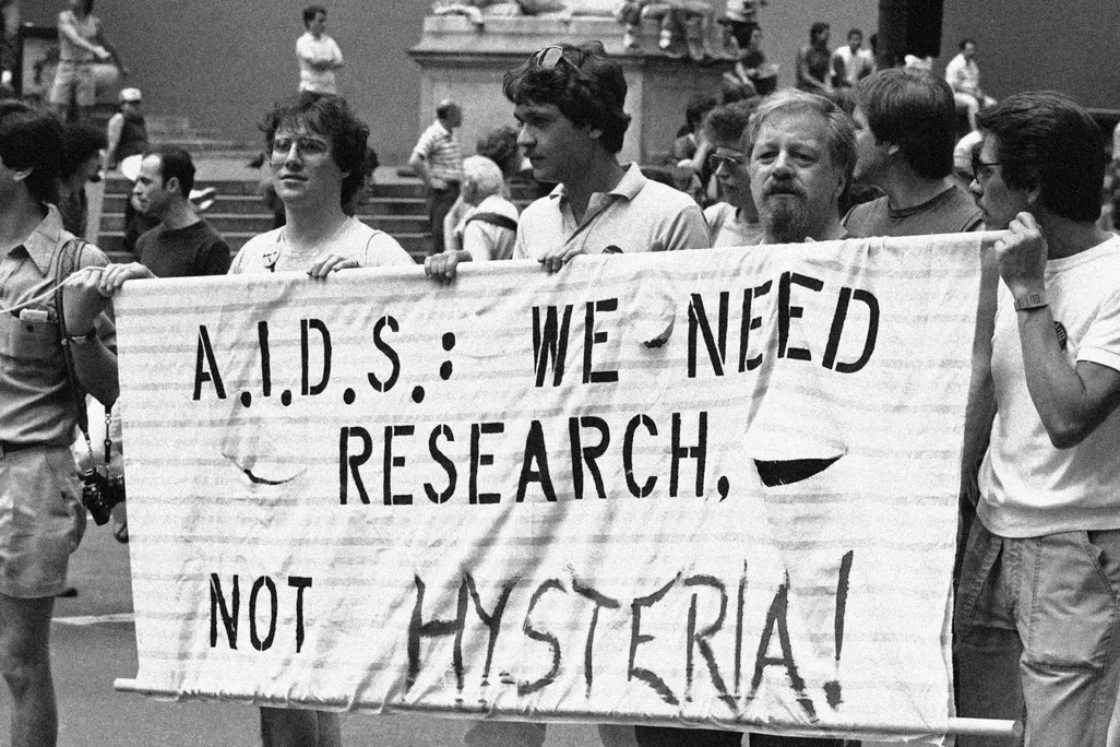 Black-and-white photograph of a group of protesters in an outdoor public space holding a large hand-painted banner that reads 'A.I.D.S.: WE NEED RESEARCH, NOT HYSTERIA!'