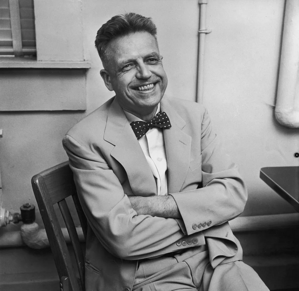 Black-and-white photograph of Alfred Kinsey, a middle-aged man with a broad, warm smile, wearing a light-colored suit and a polka-dot bow tie. He sits in a chair with his arms crossed, appearing relaxed and approachable.