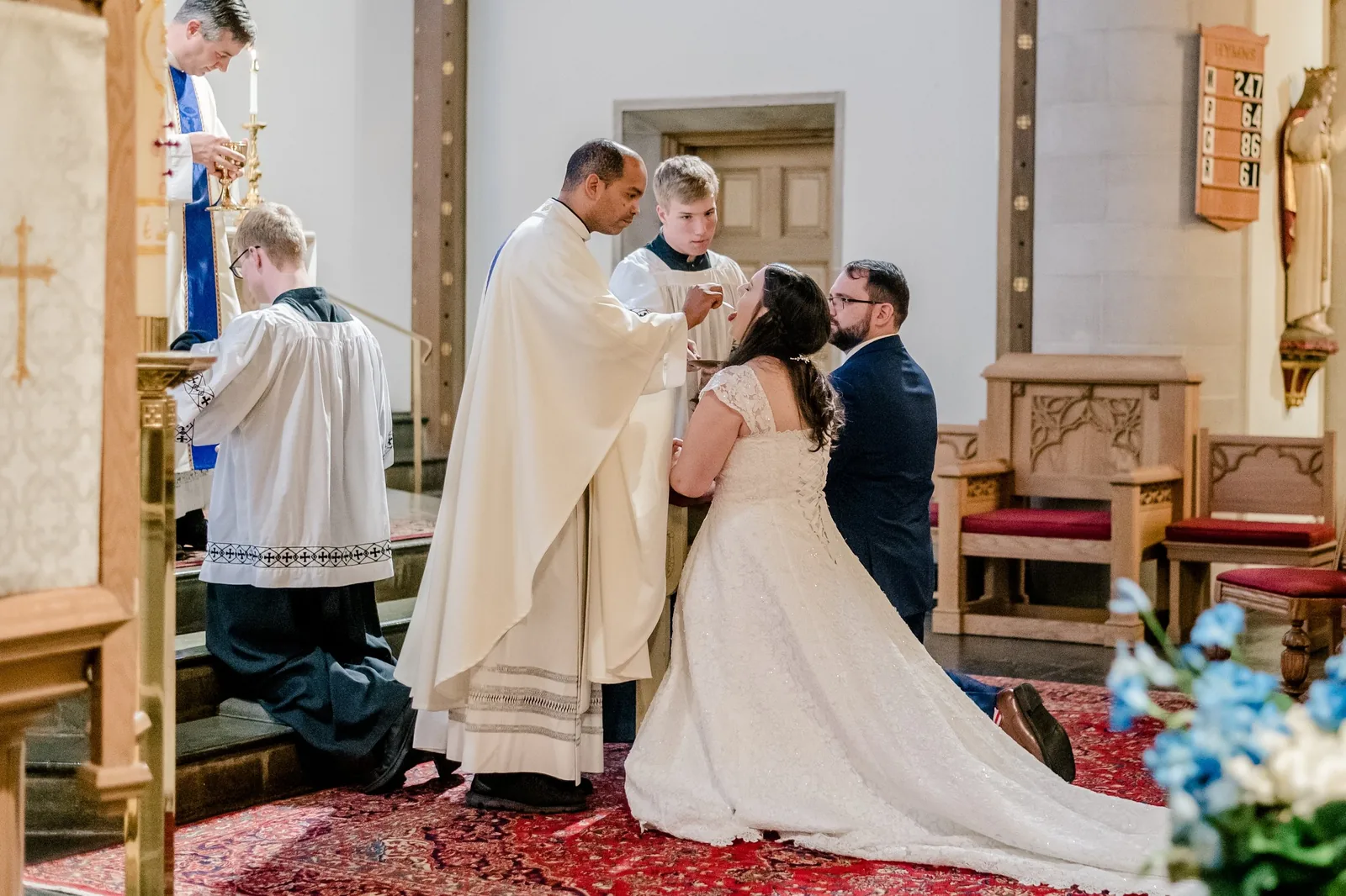 Color photograph of a Catholic wedding ceremony inside a church. The bride in a white lace gown and the groom in a dark suit kneel at the altar while a priest in white vestments administers a blessing. An altar server stands nearby, with church furnishings and floral decorations visible in the background.
