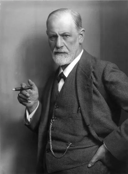 Black-and-white portrait photograph of Sigmund Freud, an older man with a neatly trimmed beard, wearing a three-piece suit and tie. He holds a cigar in his right hand, has a pocket watch chain across his vest, and gazes directly at the camera with a serious, composed expression.