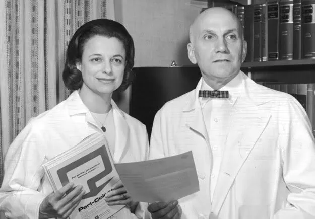 Black-and-white photograph of Virginia Johnson (left) and William Masters (right), both wearing white laboratory coats and holding documents or journals. They stand side by side before a bookshelf, looking at the camera.