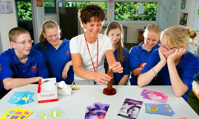 Color photograph of a female teacher leaning across a table, engaged with a group of five teenagers in matching blue school uniforms. Several colorful books and sexual health educational materials are spread on the table in a bright classroom setting.
