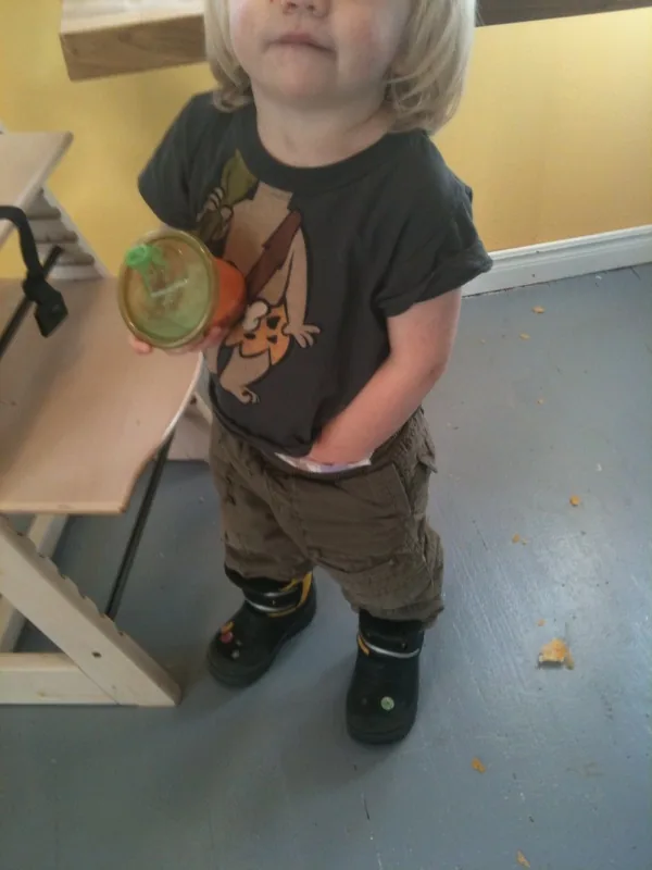A young toddler standing indoors, holding a sippy cup and exploring the world through touch.