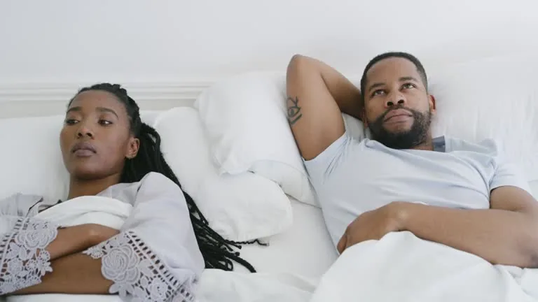 A couple sitting apart on a couch, appearing bored and disconnected.