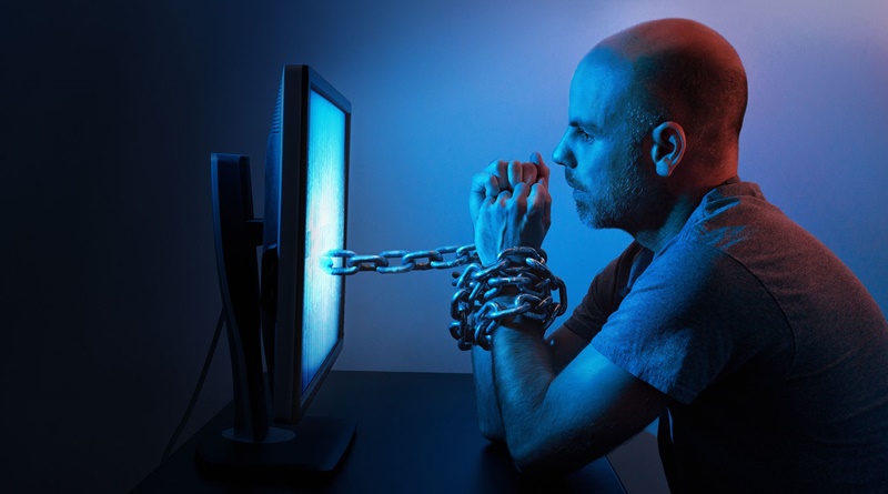 Man in dim blue light with wrists chained to a glowing computer monitor, illustrating compulsive screen use.