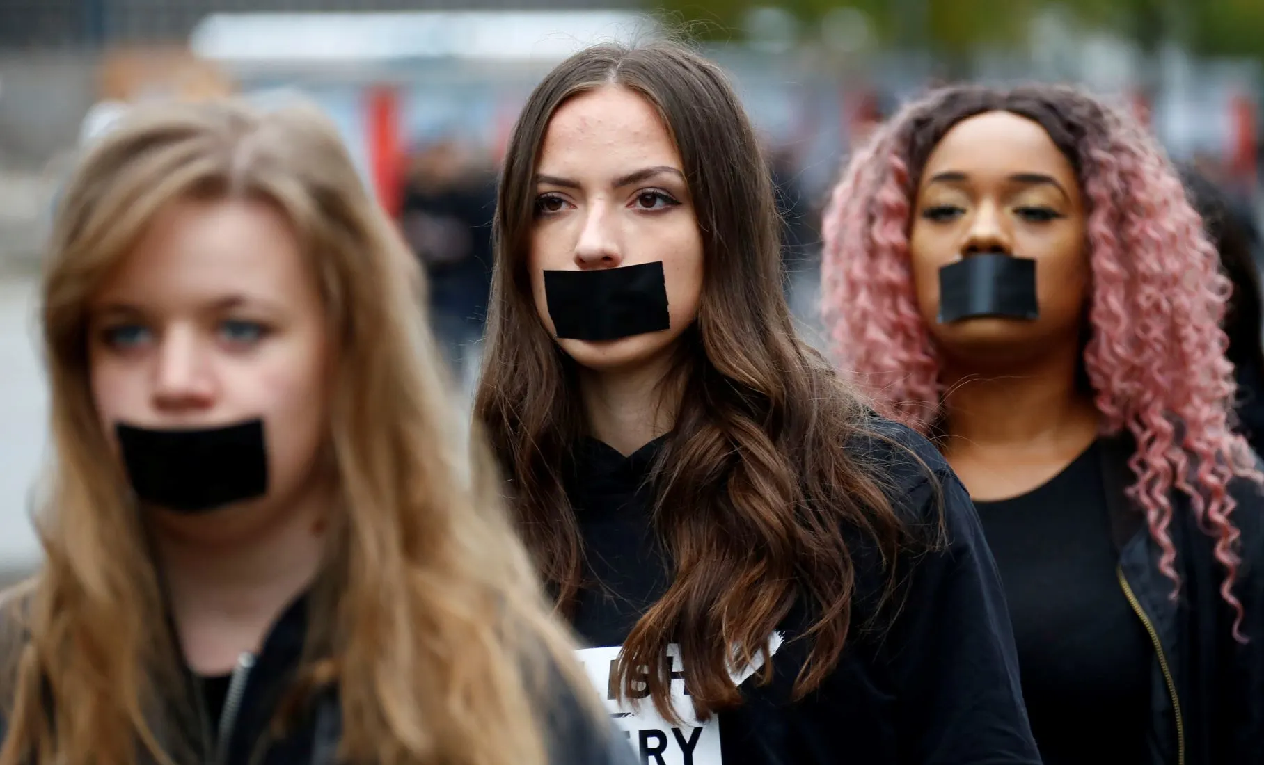 Three young people at a protest with black tape covering their mouths, symbolizing silenced victims of trafficking.