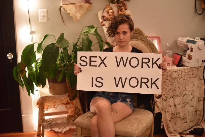 Clothed person seated in a chair holding a sign that reads SEX WORK IS WORK in a home setting.