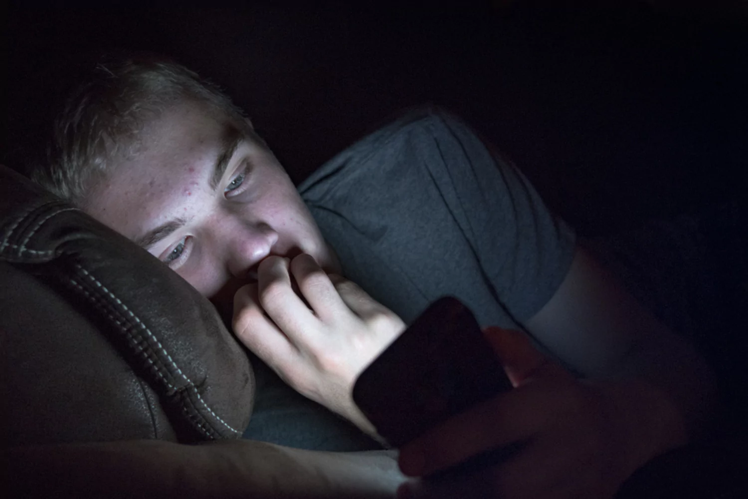 Young man lying in the dark, face illuminated by the glow of a smartphone he is looking at.