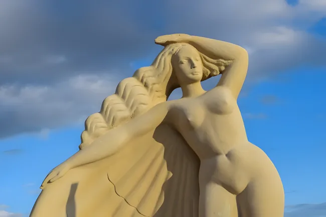 Close-up photograph of a stone sculpture of a nude female figure, shown from approximately the torso up, with one arm raised above the head. Photographed against a blue sky with clouds.