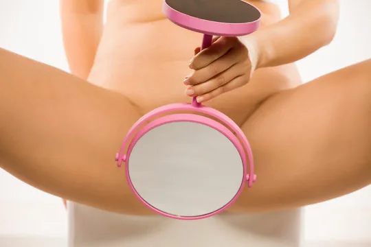 Photograph of a person, shown from the waist down with legs apart, holding a round pink-handled mirror positioned between their legs, illustrating the practice of vulvar self-examination.